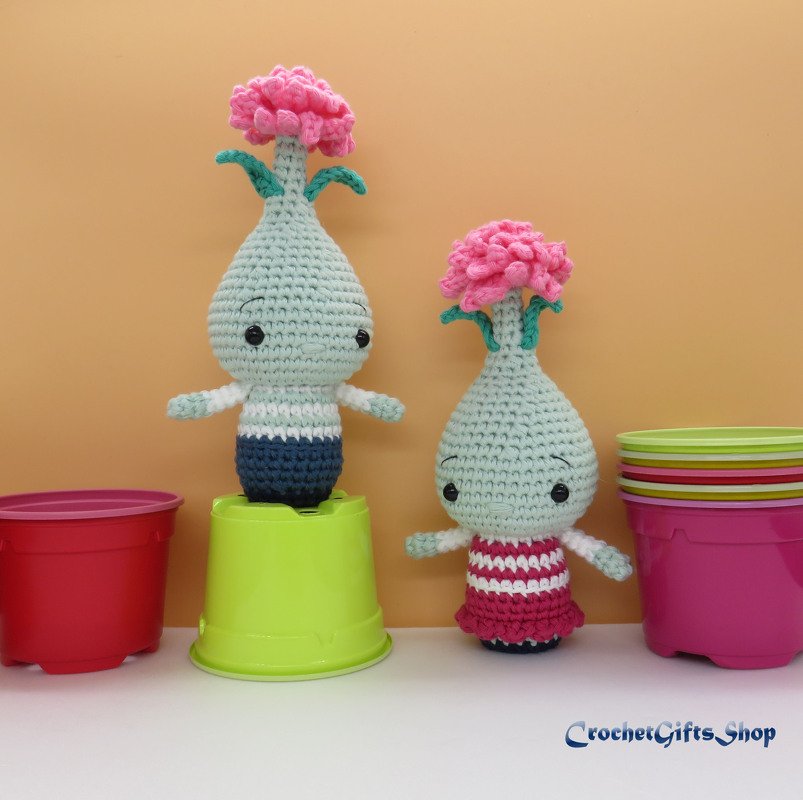Two amigurumi crochet plant dolls with pink flowers, one in blue stripes and one in pink, positioned on inverted colorful pots against a plain background.