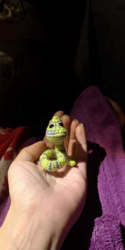 Winzige gehäkelte Schlange in Grün und Grau mit geringeltem Körper, gehalten in einer Hand.