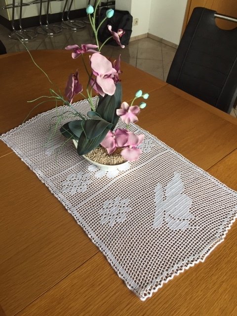 Weißer gehäkelter Tischläufer auf Holztisch mit rosa-lila Orchidee und Hasenmuster