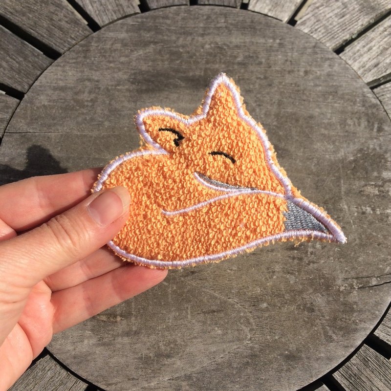 Hand hält orangefarbene Frottee-Applikation in Form eines lächelnden Fuchskopfes auf einem Holztisch