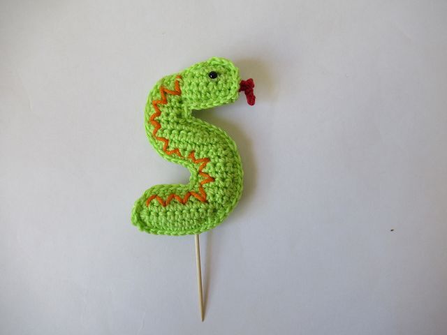 Hellgrüne gehäkelte Schlange mit orangefarbenem Zickzackmuster und Holzspieß auf weißem Hintergrund