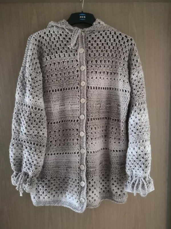 Hellgrauer gehäkelter Cardigan mit Kapuze, Knöpfen und Rüschen an den Ärmeln an einem Kleiderbügel.