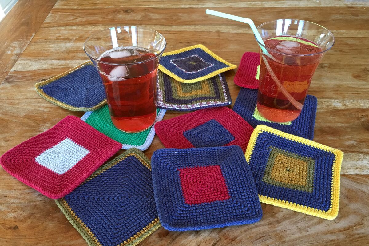 Mehrere bunte, quadratische gehäkelte Untersetzer aus Garn auf einem Holztisch, zwei Gläser mit rotem Getränk darauf