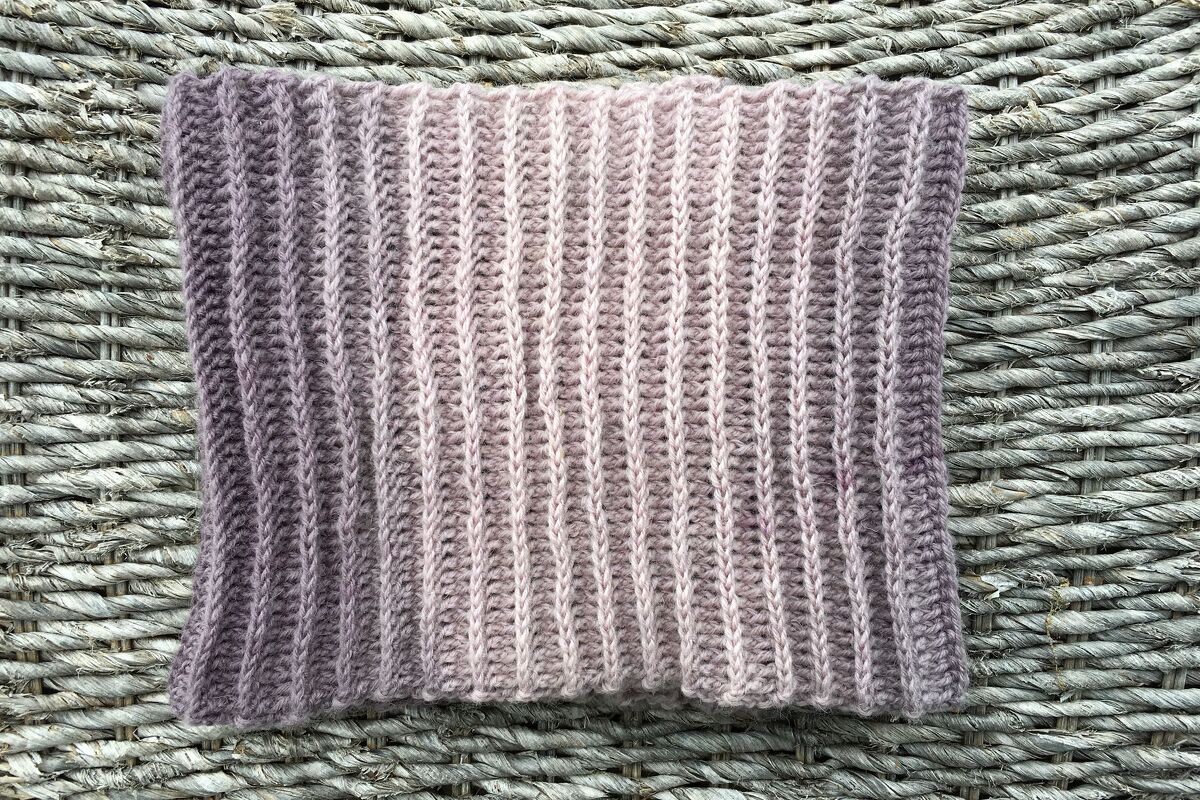 Gehäkelter Loop‑Schal mit vertikalem Rippenmuster und Lila‑Rosa‑Farbverlauf auf geflochtenem Untergrund