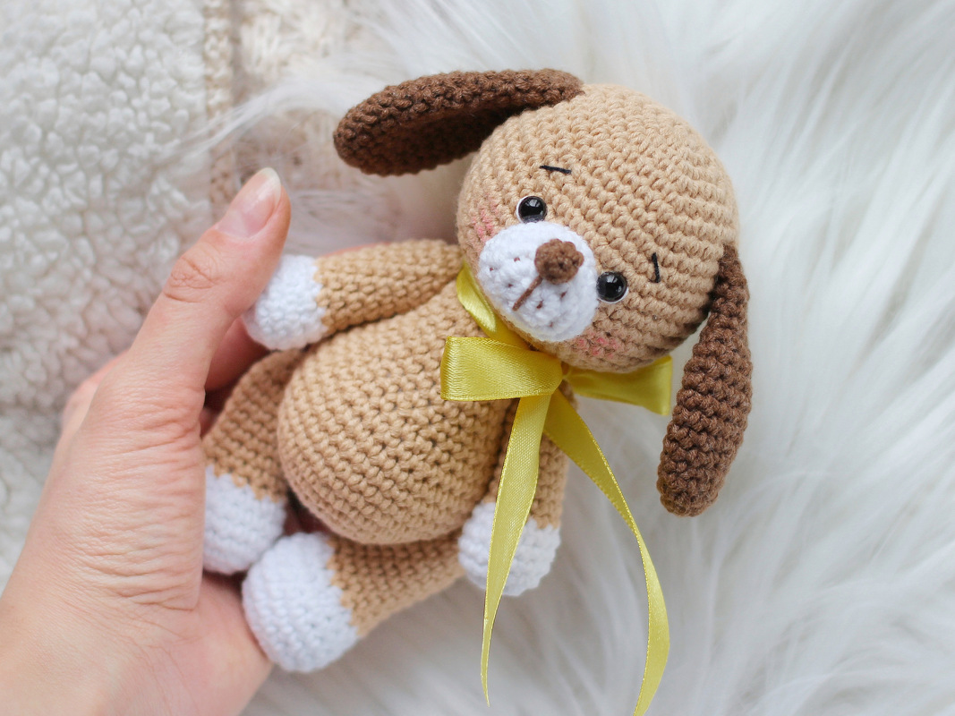 Close-up of a small brown crocheted amigurumi puppy with white paws, dark brown ears, and a yellow ribbon held in a hand