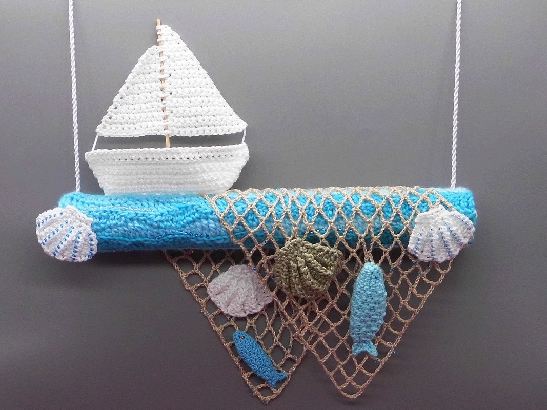 Gehäkelte maritime Wanddekoration mit weißem Segelboot auf blauem gerolltem Untergrund, beigefarbenem Fischernetz sowie gehäkelten Muscheln und Fischen vor grauer Wand.