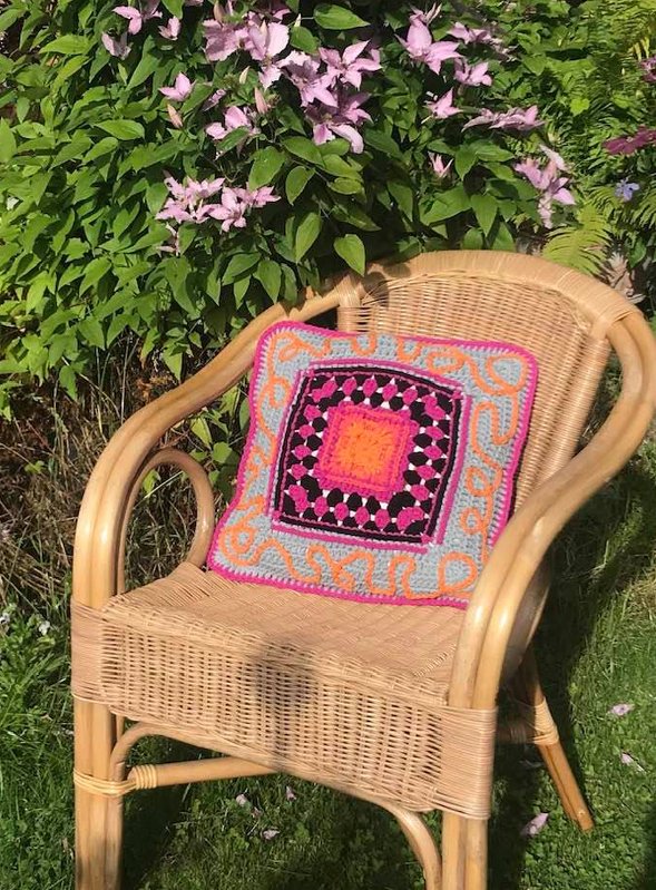 Handgehäkeltes quadratisches Kissen mit buntem Granny-Square-Muster auf einem Korbstuhl vor grünen Pflanzen und rosa Blüten.