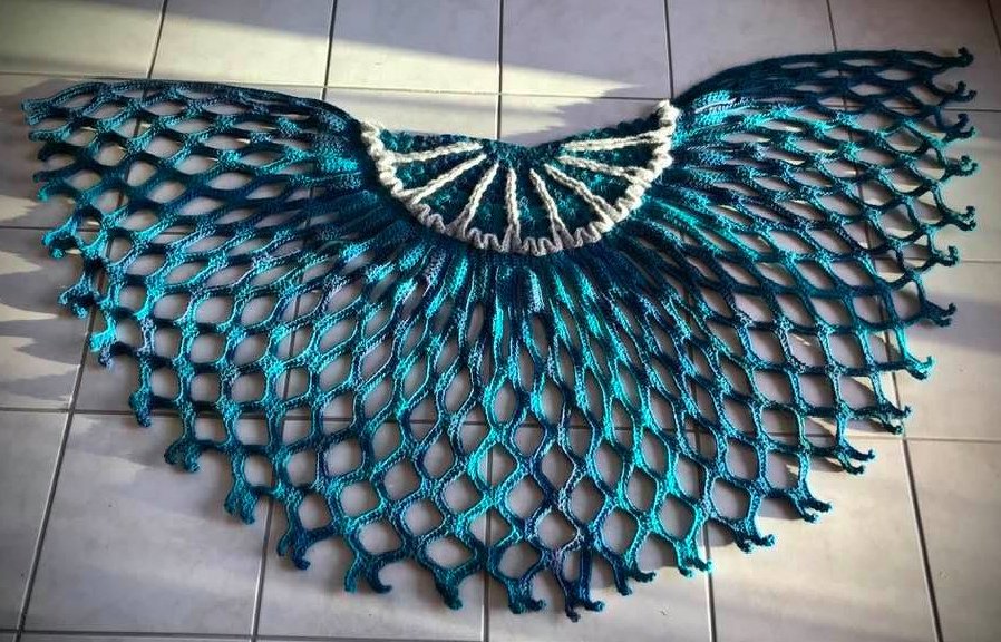 Halbrundes, gehäkeltes Tuch in Blau- und Türkistönen mit offenem Netzmuster und hellem Rand, auf hellen Bodenfliesen gelegt.