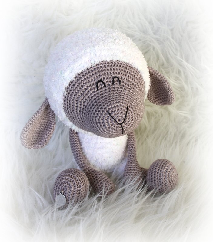 Gehäkeltes Amigurumi-Schaf mit weißem flauschigem Kopf und beige-braunem Gesicht, sitzend auf hellem Kunstfell