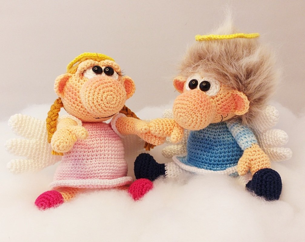 Two crocheted amigurumi angels holding hands — pink dress with braids and blue outfit with fluffy yarn hair, both with wings and yellow halos