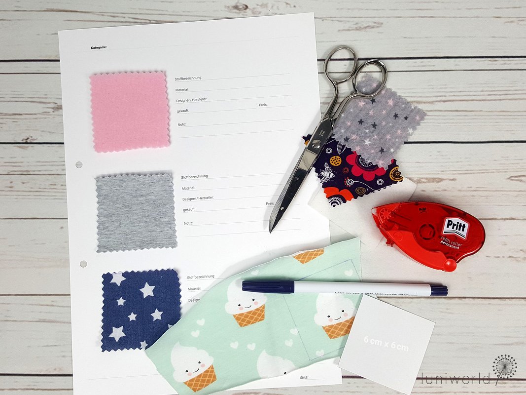 Nahaufnahme eines Stoffpasses mit rosa-, grau- und blaugemusterten Stoffmustern, umgeben von einer Schere, einem Kleberoller, einem Stift und weiteren Stoffresten auf einem Holztisch.