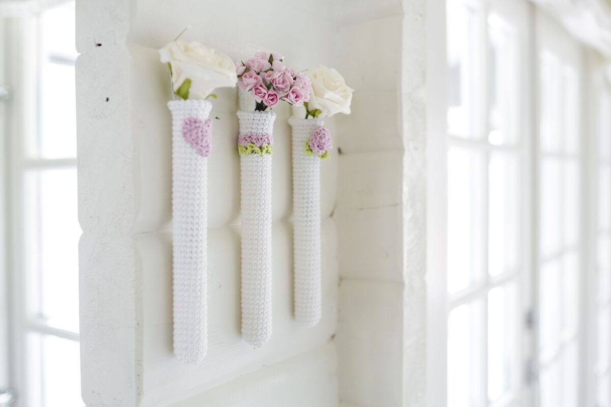 Drei weiße, gehäkelte Röhrenvasen mit rosa und weißen Blüten an einer weißen Holztafel