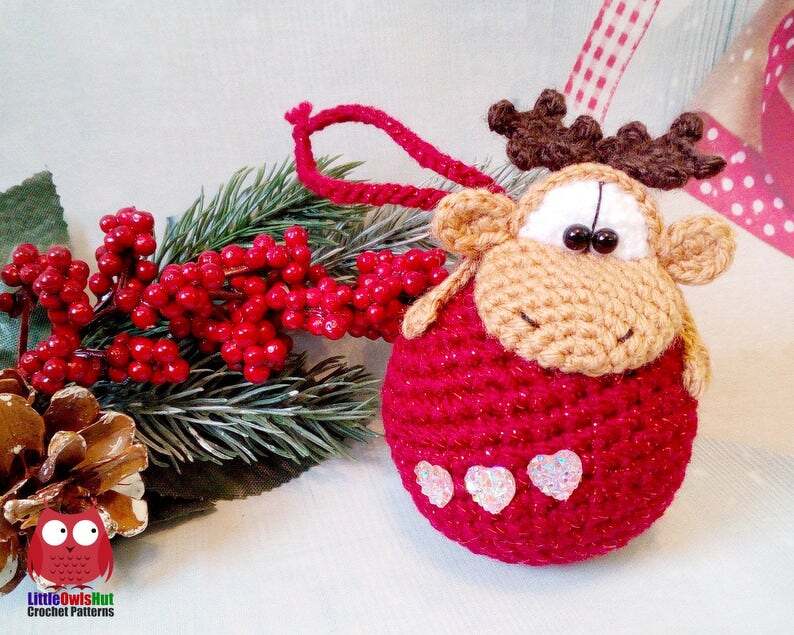 Crocheted reindeer ornament with a red round body, beige face, brown antlers and three iridescent heart appliqués, shown with red berries and an evergreen sprig.
