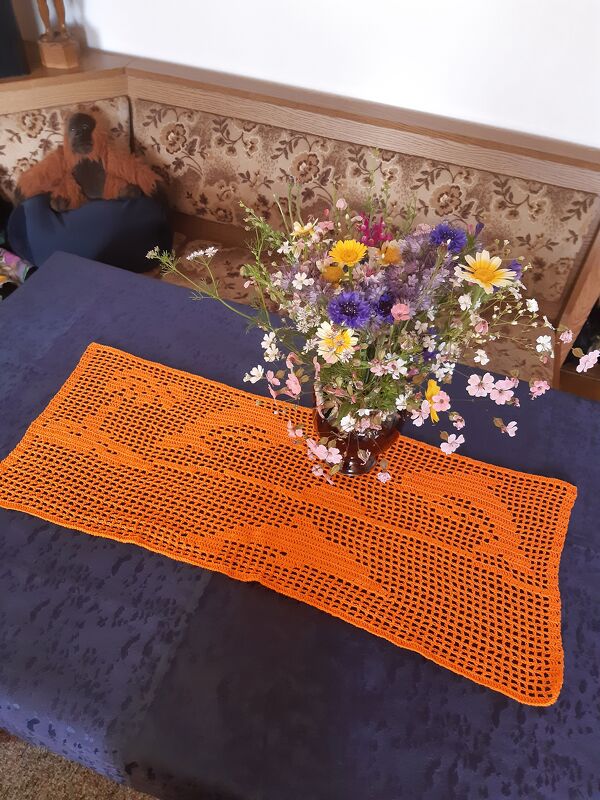 Orangefarbener gehäkelter Tischläufer im Filet-Häkelmuster auf dunkler Tischdecke mit Vase und Wiesenblumen
