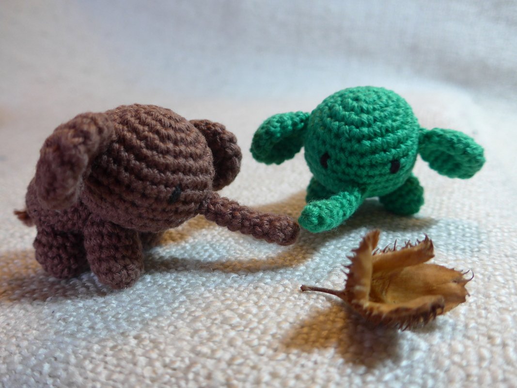 Zwei gehäkelte Amigurumi-Elefanten (braun und grün) auf hellem Stoff neben einem getrockneten Blatt