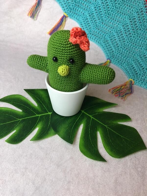Grüner Amigurumi-Häkelkaktus mit orangefarbener Blüte im weißen Topf auf Monstera-Blatt