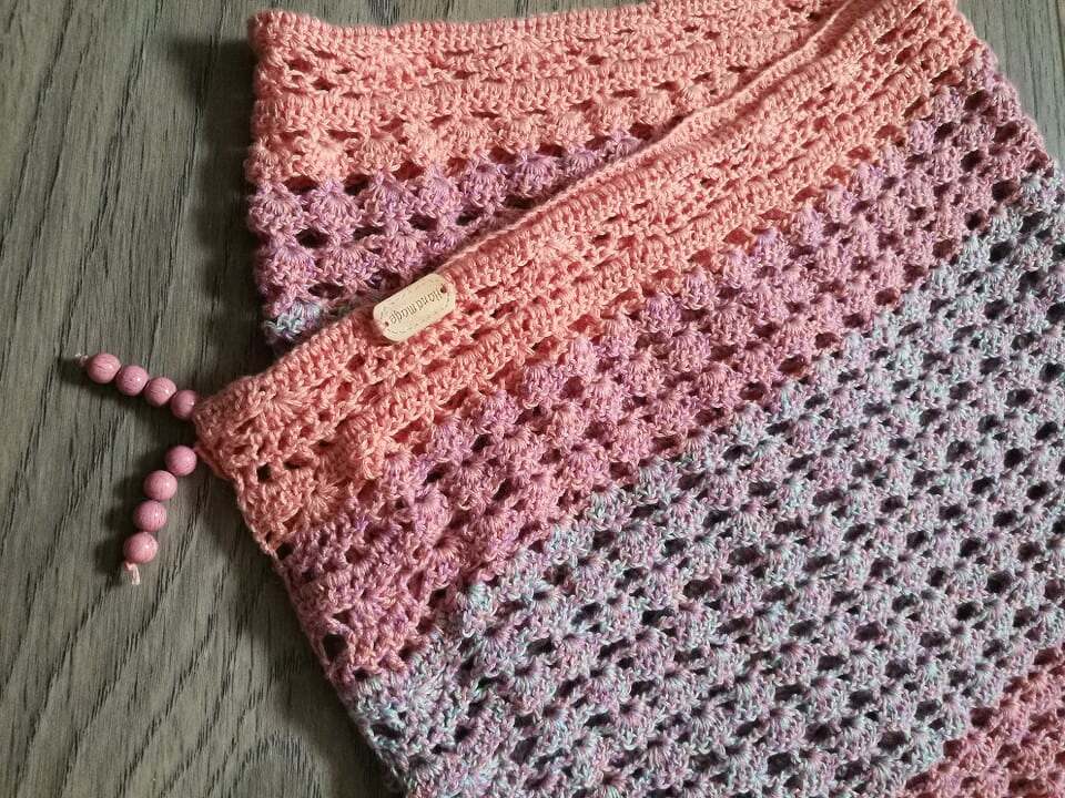 Handgehäkelter Schal mit Ombé-Verlauf (Rosa–Lila–Hellblau), offenes Maschenmuster, Holzperlen und kleines Etikett auf Holzuntergrund.