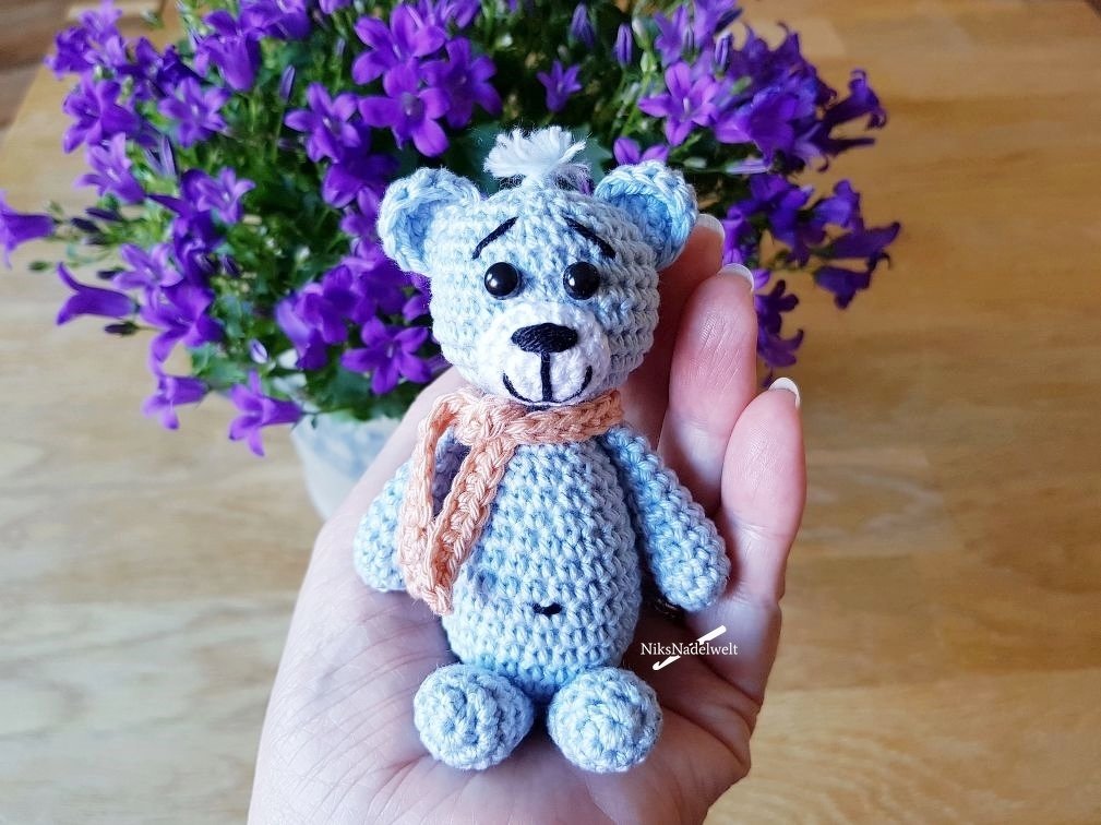 Hellblauer gehäkelter Mini-Teddybär mit Schal, in einer Hand gehalten, vor violetten Blüten