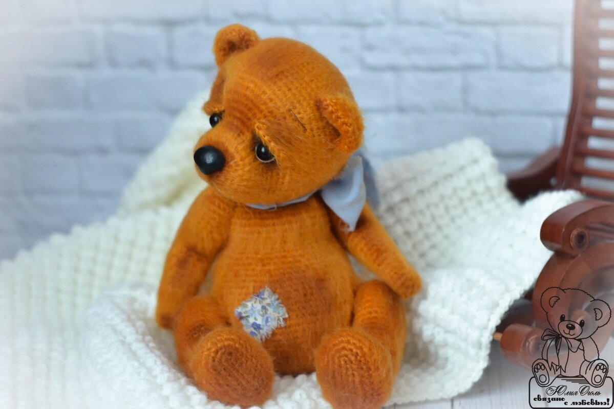 Gehäkelter orangebrauner Teddybär mit hellblauem Halstuch und floralem Patch auf einer weißen Decke vor einer Ziegelwand.