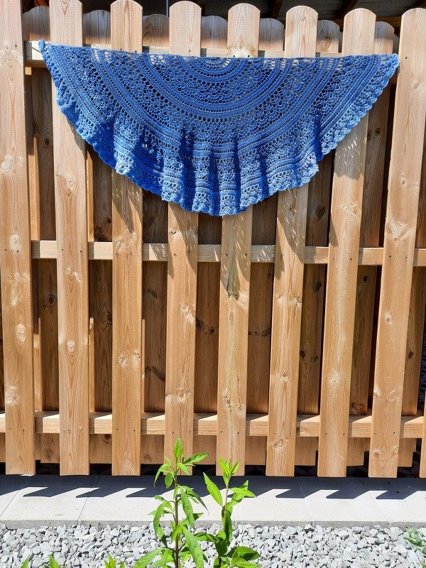 Blue crocheted crescent shawl with lace stitches draped on a wooden fence.