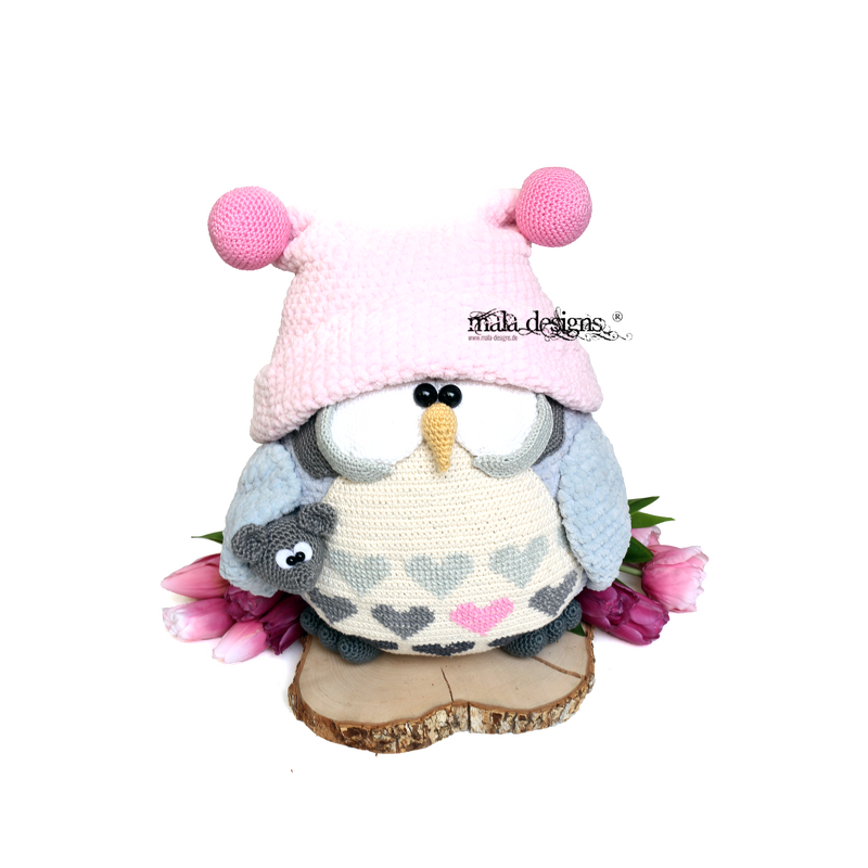 Crocheted amigurumi owl wearing a pink bobble hat, heart-patterned belly, holding a small grey amigurumi