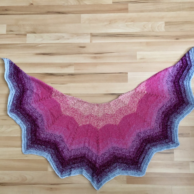 Halbrundes gestricktes Tuch mit Farbverlauf von Pink zu Blau auf hellem Holzboden