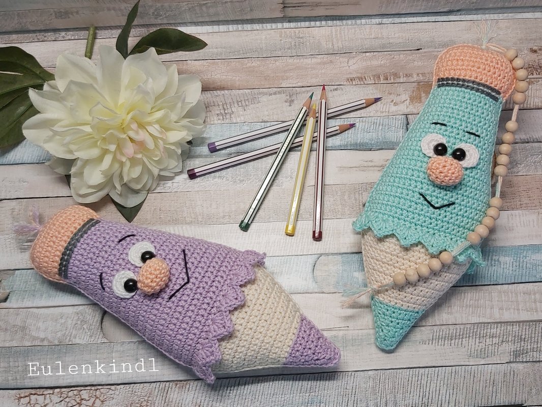 Zwei gehäkelte Stiftfiguren mit Augen und Nase, auf hellem Holzboden liegend. Ein türkisfarbener und ein lila Stift mit gestreiften Spitzen und holzfarbenen Abschnitten.