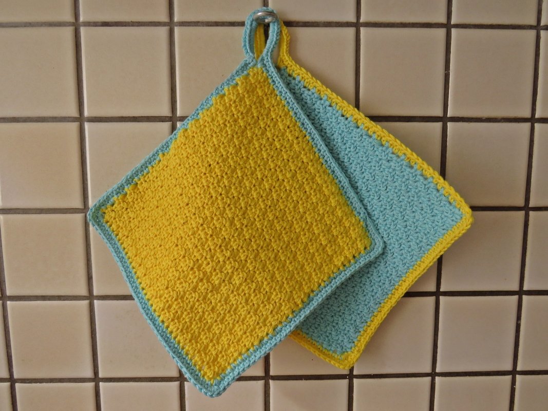 Zwei gehäkelte Topflappen in Gelb und Hellblau an einem Haken vor einer Fliesenwand