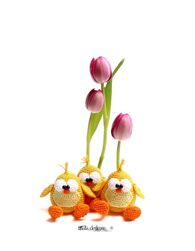 Drei gelbe gehäkelte Amigurumi-Küken vor drei rosa Tulpen auf weißem Hintergrund.