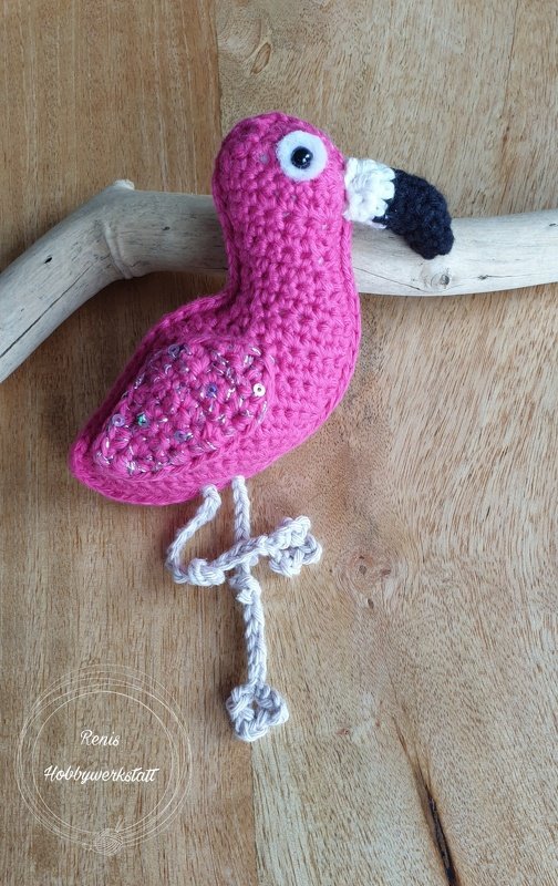 Gehäkelter pinker Flamingo-Amigurumi mit Pailletten auf den Flügeln und schwarzem Schnabel auf Holzuntergrund
