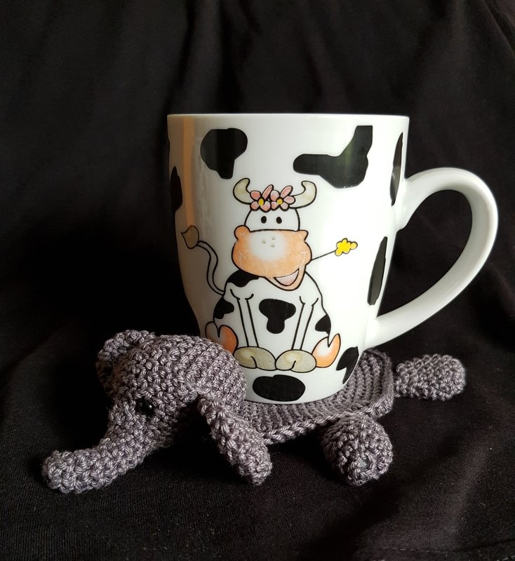Grauer, gehäkelter Elefanten-Untersetzer unter einer weißen Tasse mit Kuhmotiv auf dunklem Hintergrund.