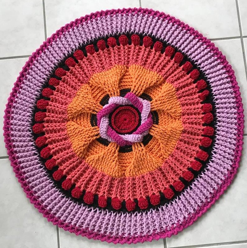 Runder gehäkelter Teppich mit Mandala-Muster in Orange, Rosa, Magenta und Schwarz auf hellem Fliesenboden