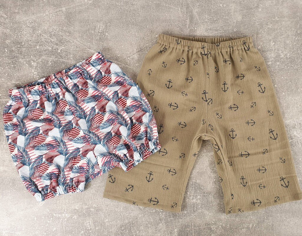 Zwei Babyhosen auf grauem Hintergrund: helle Windelhose mit buntem Blattmuster und beigefarbene Shorts mit Anker-Motiv.