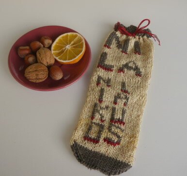 Beiger gestrickter Beutel mit roten Details und Kordel neben einer kleinen Schale mit Nüssen und einer Orangenscheibe