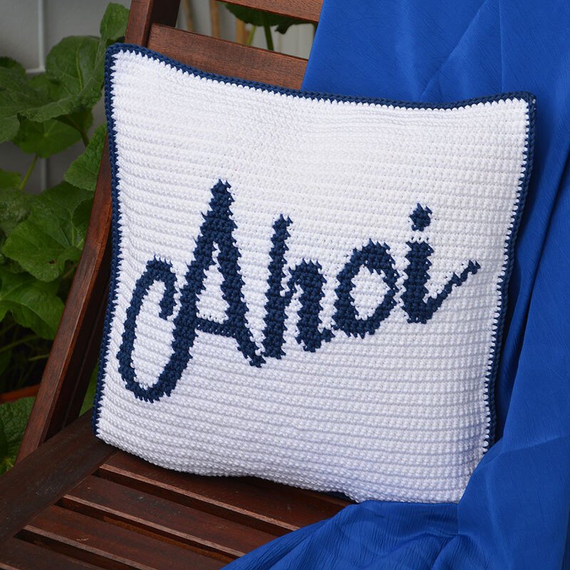 Weißes gehäkeltes Kissen mit dunkelblauem 'Ahoi'-Schriftzug auf Holzstuhl