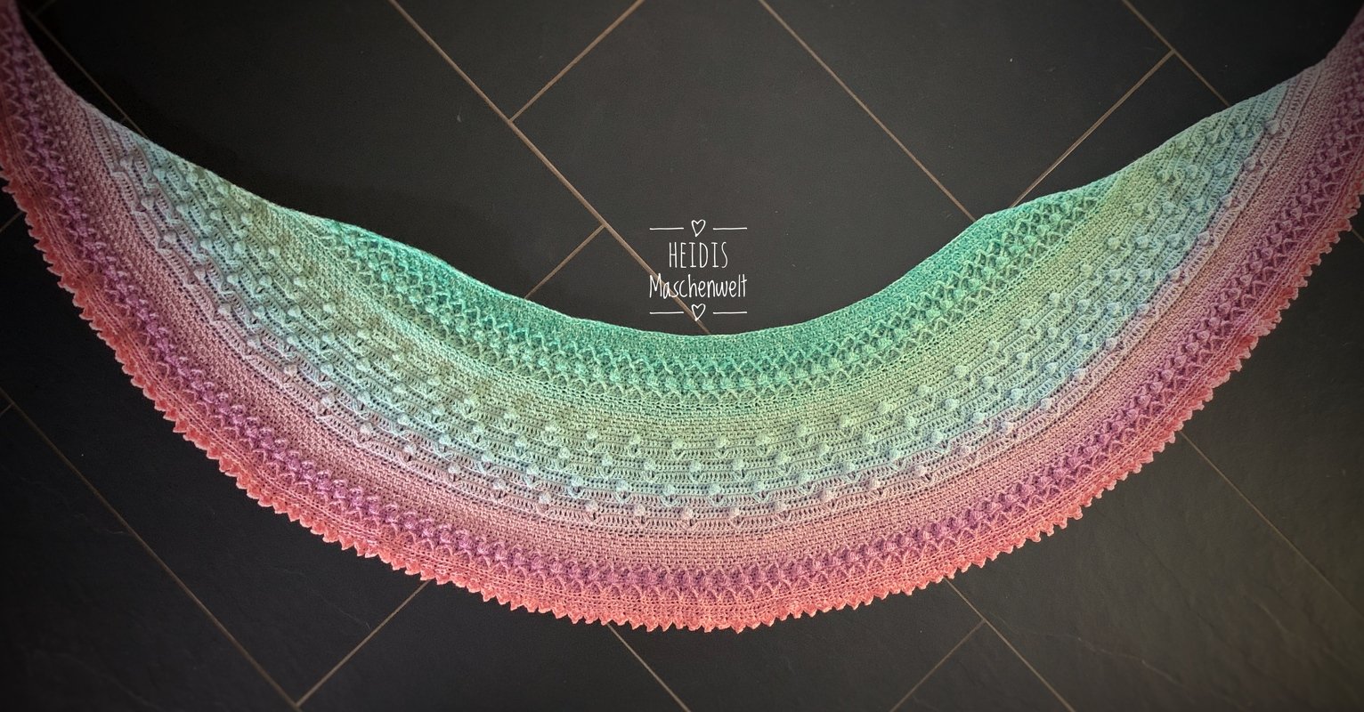 Halbrundes gehäkeltes Tuch mit Farbverlauf in Rosa, Lila, Blau und Grün auf dunklem Hintergrund.