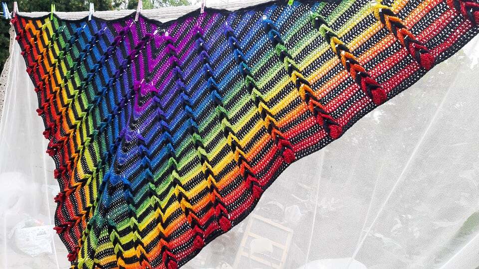 Gehäkeltes Dreieckstuch mit Zickzackmuster in Regenbogenfarben, an einer Wäscheleine im Freien aufgehängt.