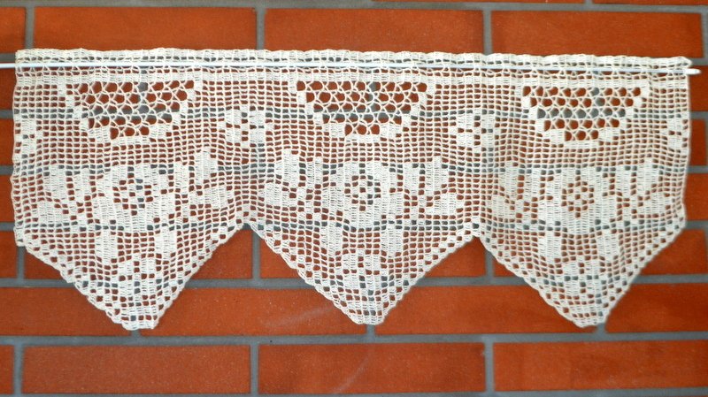 Cremefarbene gehäkelte Scheibengardine mit geometrischem Filetmuster und dreieckigem Saum vor roter Ziegelwand