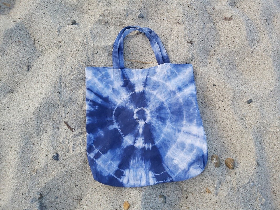 Stoff-Einkaufstasche mit blau-weißem Tie‑Dye-Spiralmuster auf Sand