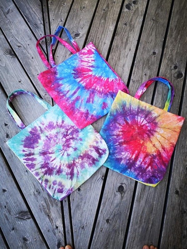 Drei bunte Tie‑Dye Stofftaschen mit Spiralmustern auf Holzdielen.