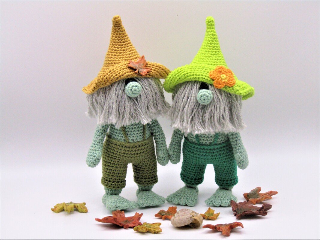 Zwei gehäkelte Zwergfiguren mit spitzen Mützen (gelb, hellgrün), grauen Bärten und grünen Overalls, vor weißen Hintergrund mit Herbstblättern.