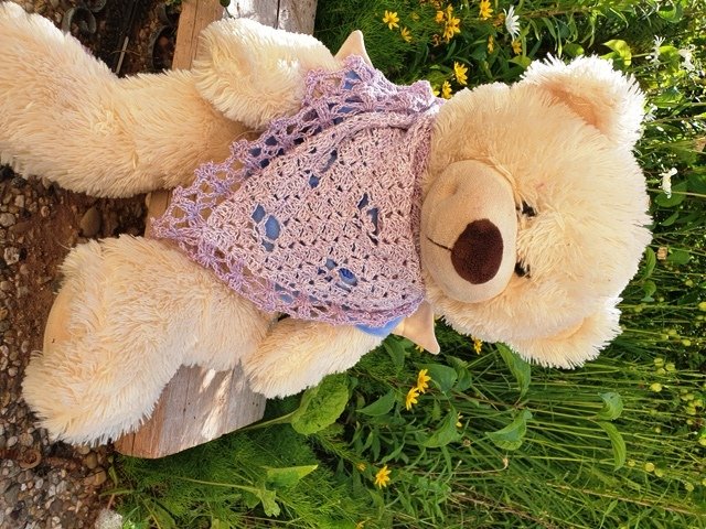 Beige teddy bear wearing a lilac lacy crocheted triangle shawl outdoors.