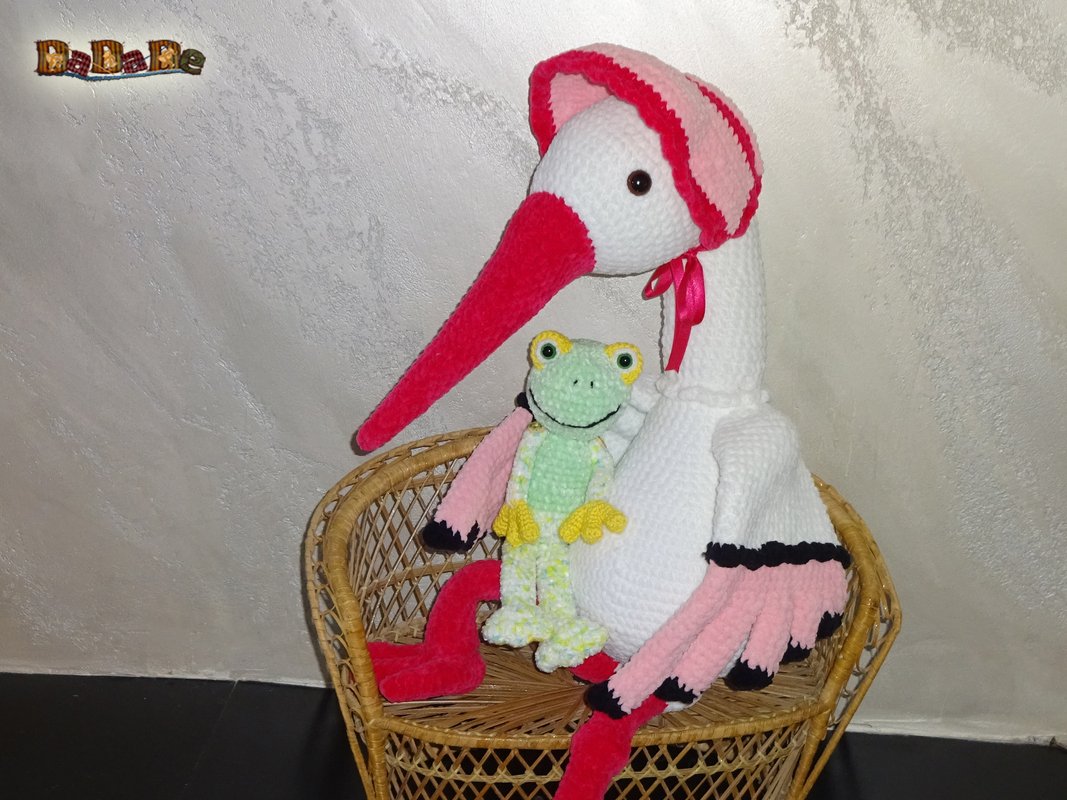 Weißer gehäkelter Storch mit rosa Haube neben einem grünen Häkelfrosch im Korbsessel.