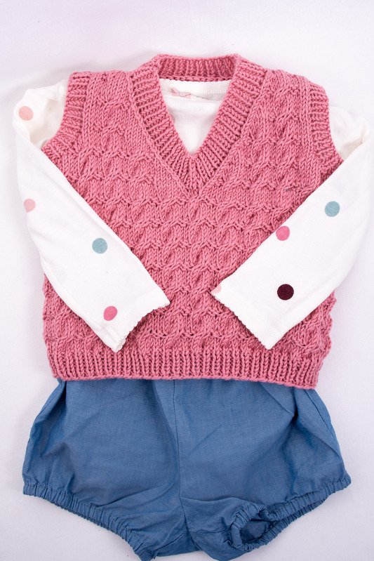 Rosa gestrickte ärmellose Babyweste mit VAusschnitt ber weißem Langarmshirt mit bunten Punkten und blauer Pumphose