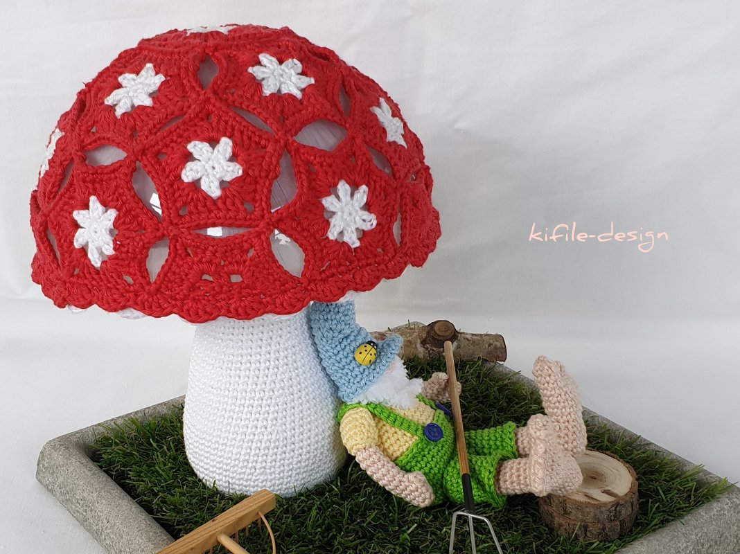 Gehäkelter roter Pilz mit weißen Sternen und liegender Wichtel auf Kunstrasen in grauer Schale