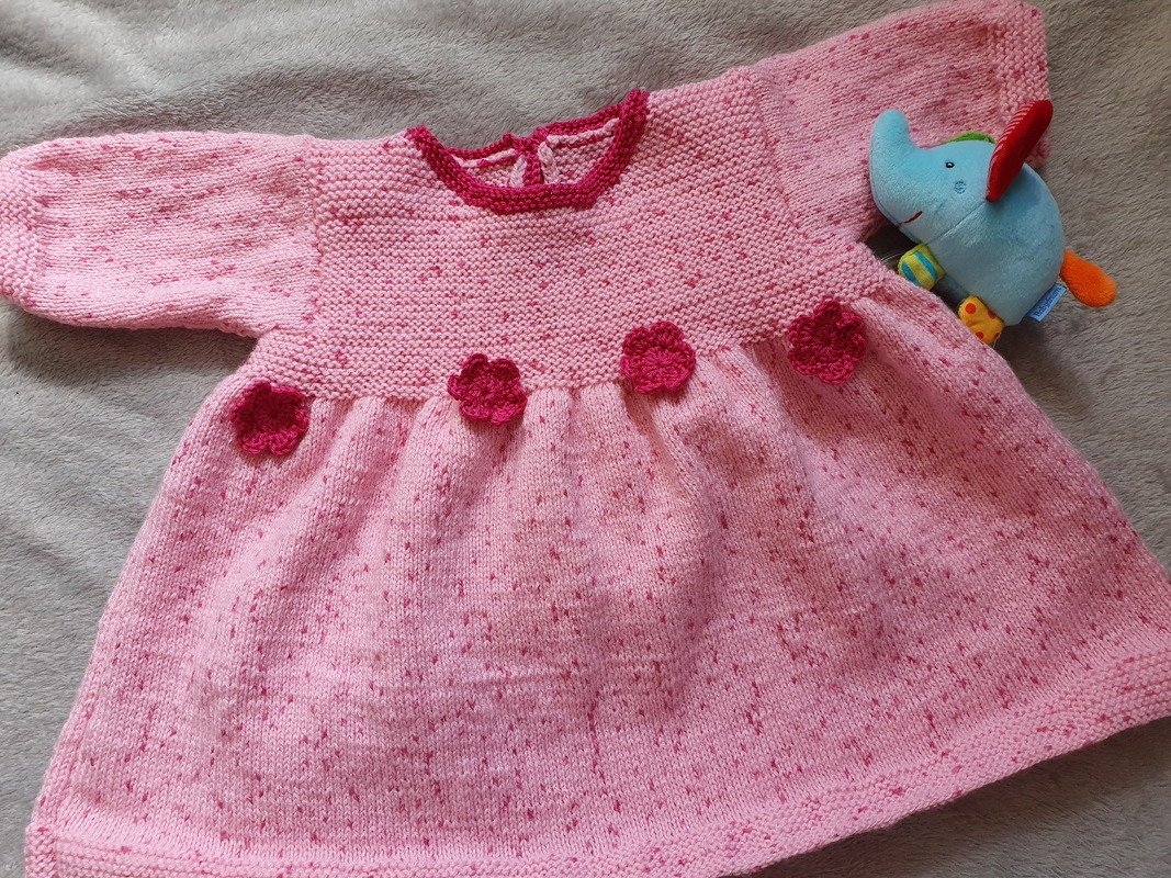 Rosa gestricktes Babykleid mit langen Ärmeln, gehäkelten dunkleren Blüten an der Taille, auf hellgrauem Untergrund liegend neben einem blauen Stoffelefanten