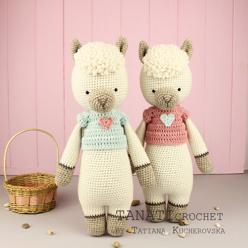 Zwei gehäkelte Amigurumi-Alpakas mit flauschiger Mähne und pastellfarbenen Shirts mit Herzapplikationen vor rosa Holzwand