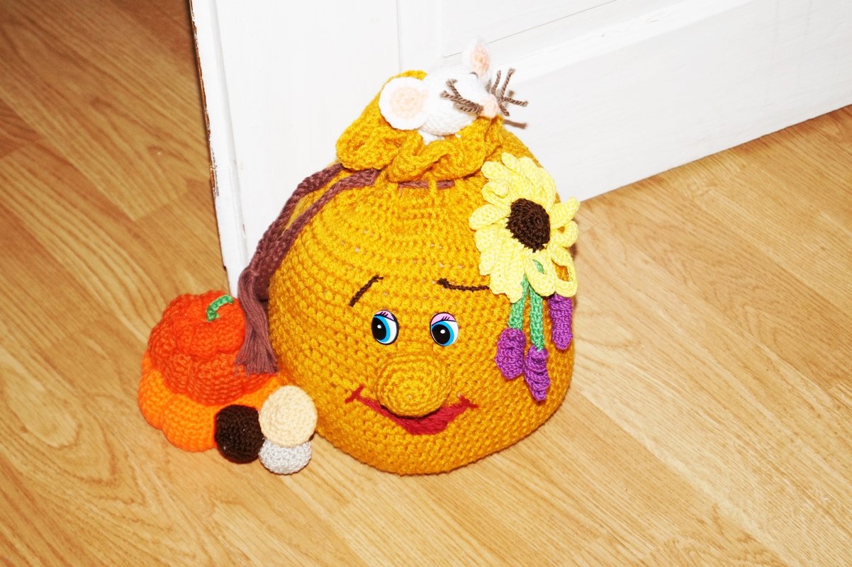 Handgehäkelter gelber Türstopper mit Gesicht, blauen Augen, Sonnenblume, lila Blüten, weißer Maus und kleinem Kürbis auf Holzfußboden