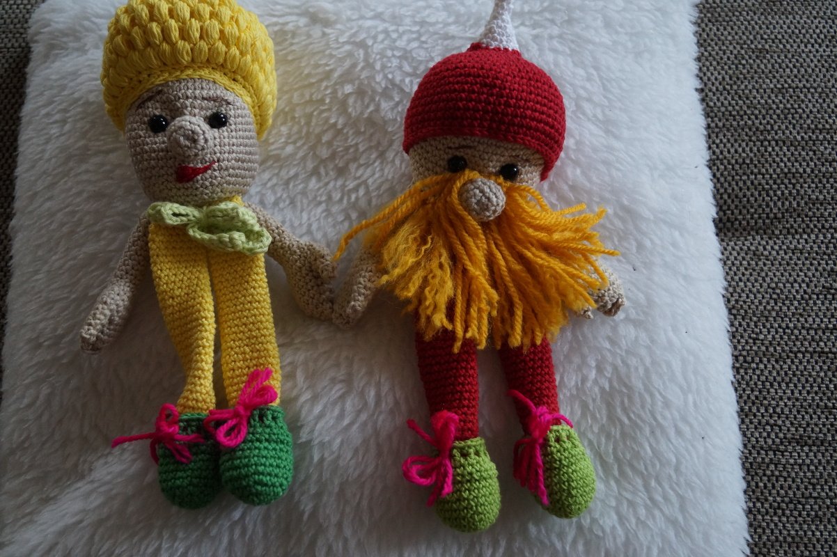 Zwei gehäkelte Amigurumi-Figuren: Elfe (gelb) und Zwerg (rot) Hand in Hand auf weißer Decke