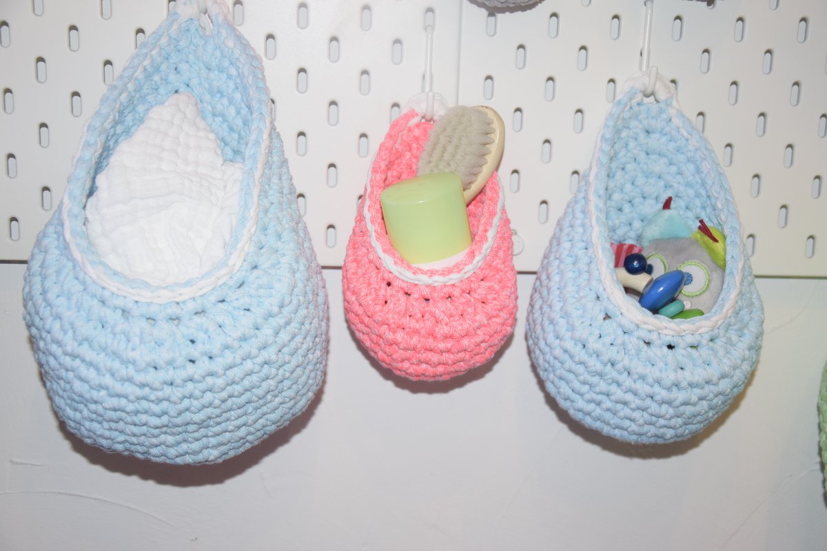 Drei gehäkelte Hängekörbe in Hellblau und Rosa an einer Lochwand, gefüllt mit Babyutensilien.